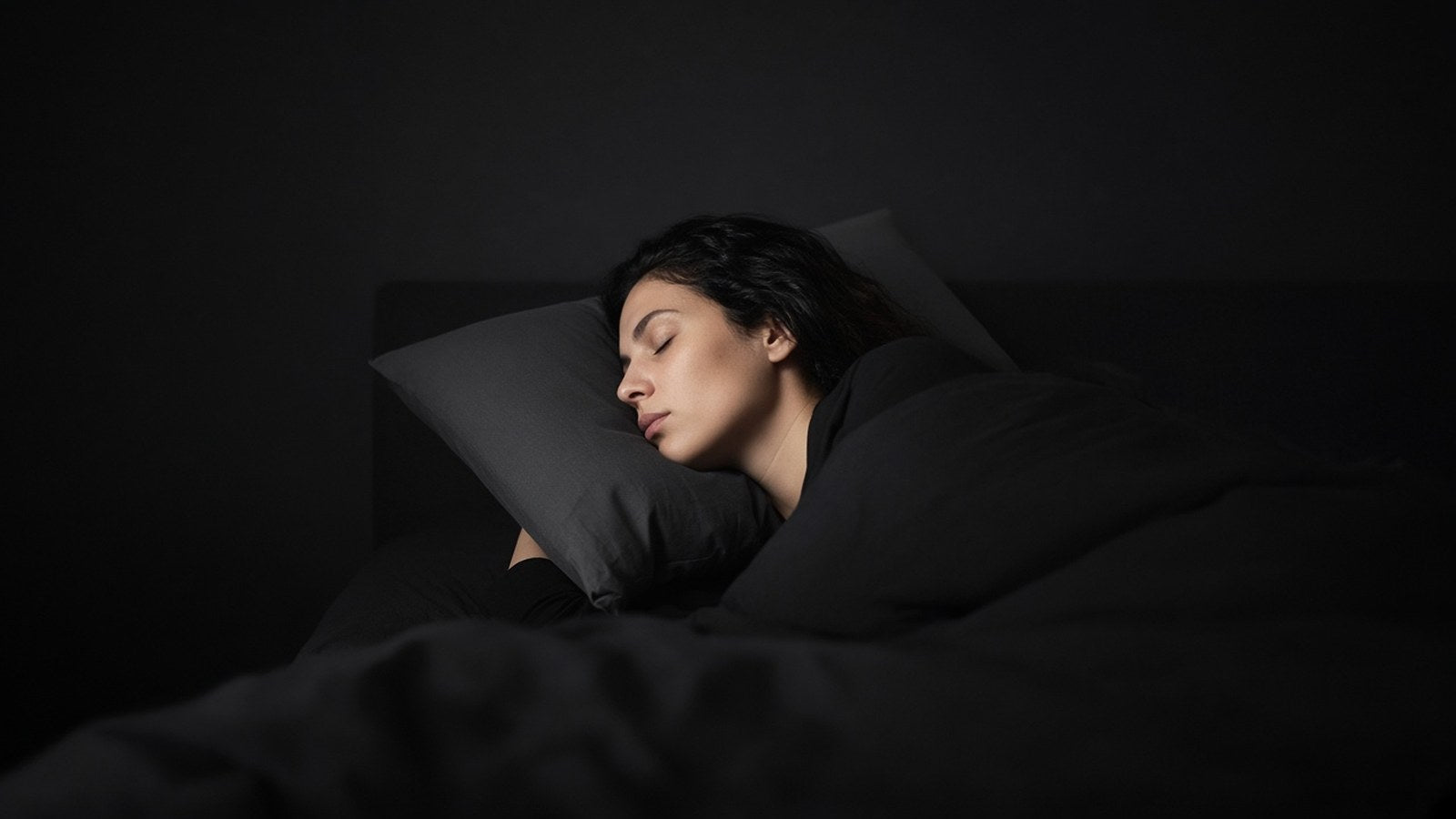 A woman sleeps on her side on a charcoal pillow in a dark bedroom, seen from the side.